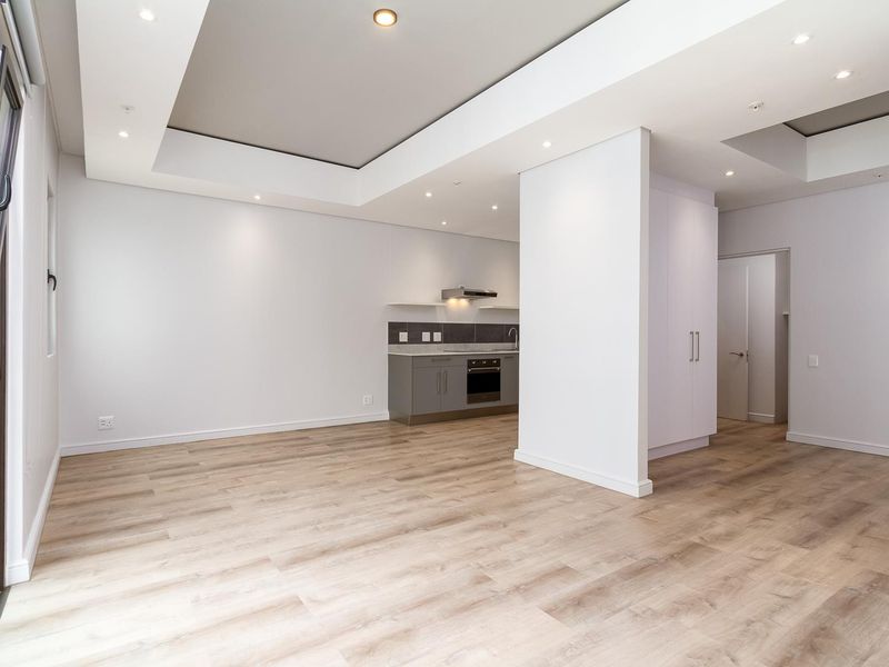 Modern open-plan living area featuring light laminate flooring, recessed ceiling lighting, and a compact kitchen with built-in cabinetry and appliances.