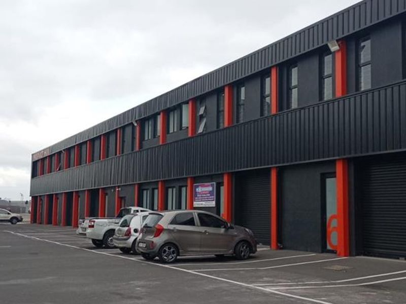 Exterior view of a modern industrial complex featuring dark metal cladding with orange accents, roller shutter doors, and a paved parking area.