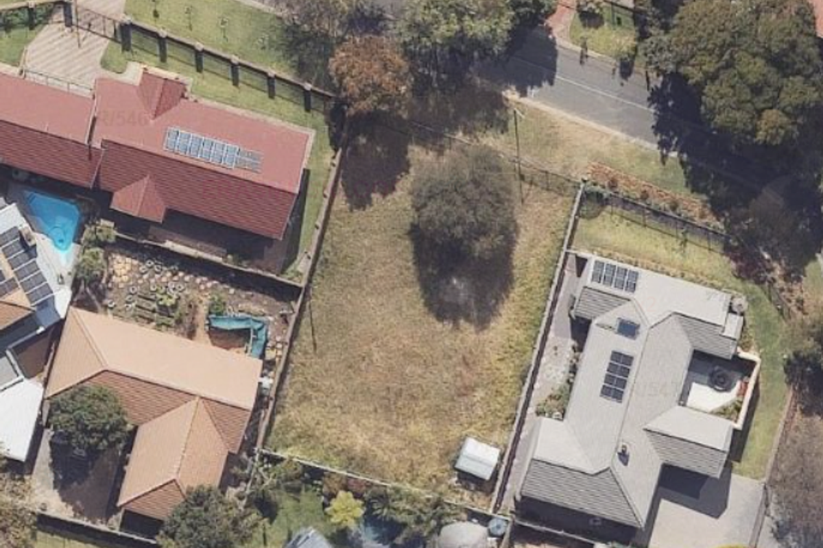 An aerial view of a vacant residential plot situated between established homes, featuring open land with some mature trees and perimeter fencing.