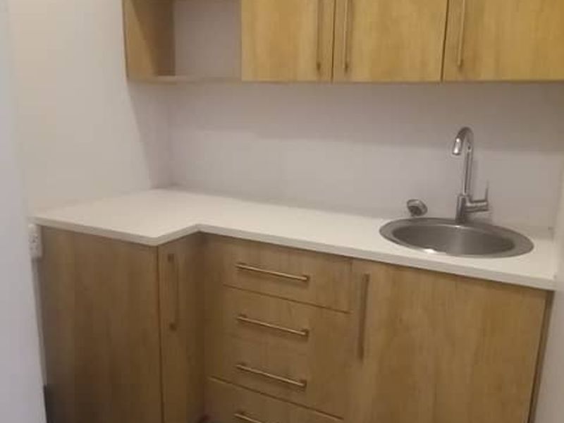 A clean and modern scullery or kitchenette area featuring light wood-finish cabinetry, a white countertop, and a stainless steel sink.