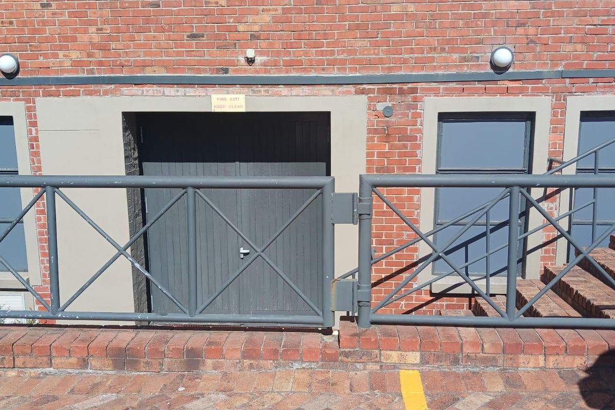 Exterior view of a brick building facade featuring a fire exit door, metal security railing, and paved ground level access.