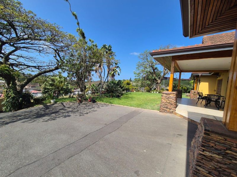 A spacious rear exterior featuring a covered patio with stone-clad pillars, overlooking a well-maintained lawn and mature garden area.