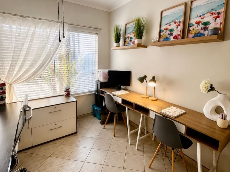A functional home office featuring tiled flooring, a long built-in desk, and large windows with venetian blinds and curtains.