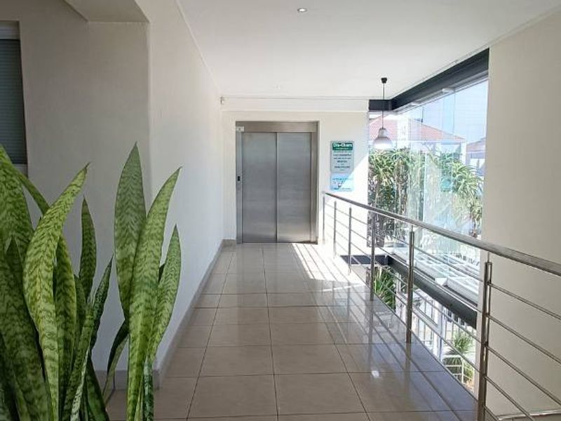 A bright, modern office building hallway featuring ceramic tiled flooring, a stainless steel balustrade, and a central elevator access point.