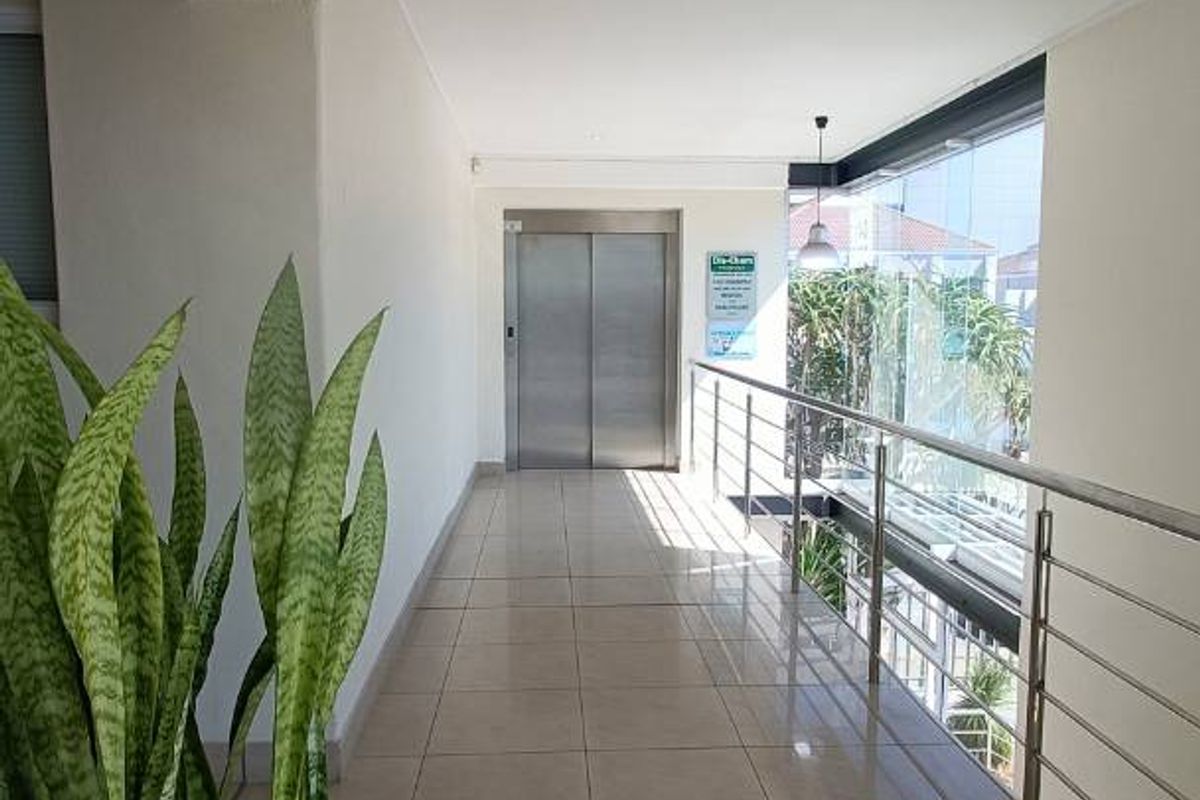 A bright, modern office building hallway featuring ceramic tiled flooring, a stainless steel balustrade, and a central elevator access point.