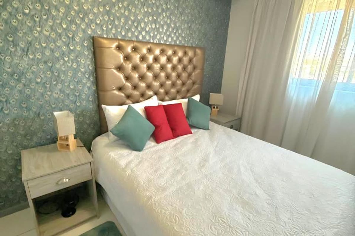 A well-maintained bedroom featuring a decorative patterned feature wall, neutral tiled flooring, and natural light through window curtains.
