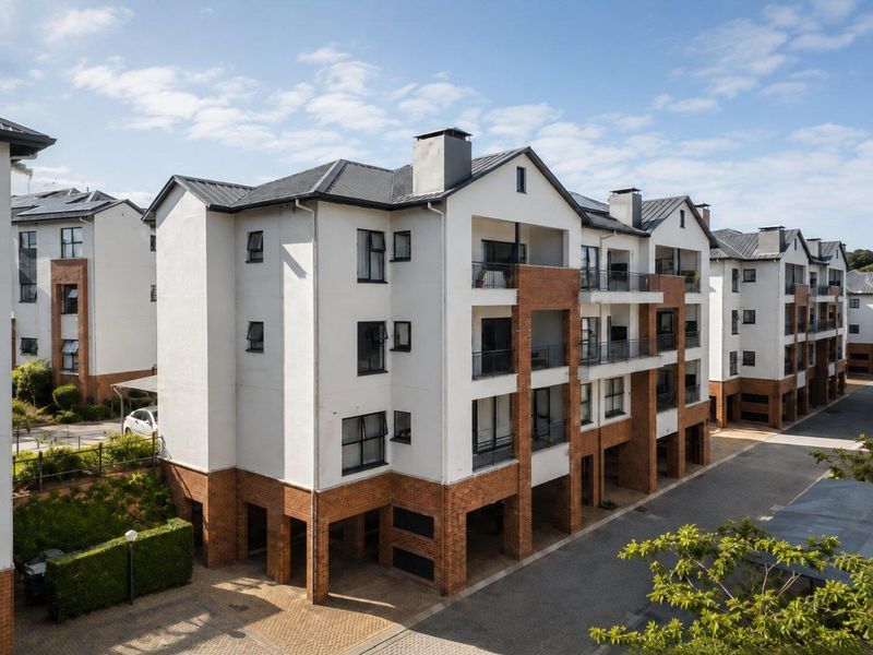 A multi-story apartment complex featuring a modern brick and plaster facade with private balconies and a paved driveway area.
