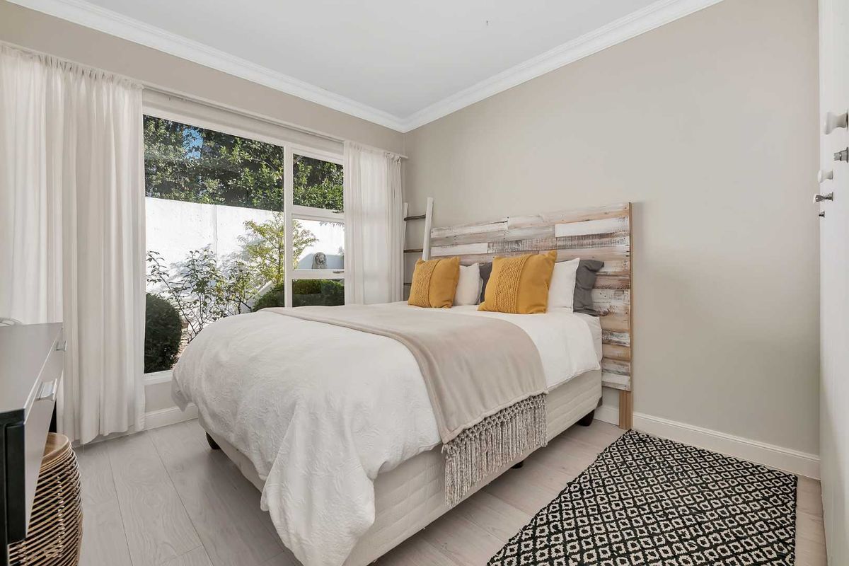 A bright and well-maintained bedroom featuring light-toned flooring, crown molding, and a large window providing natural light and garden views.