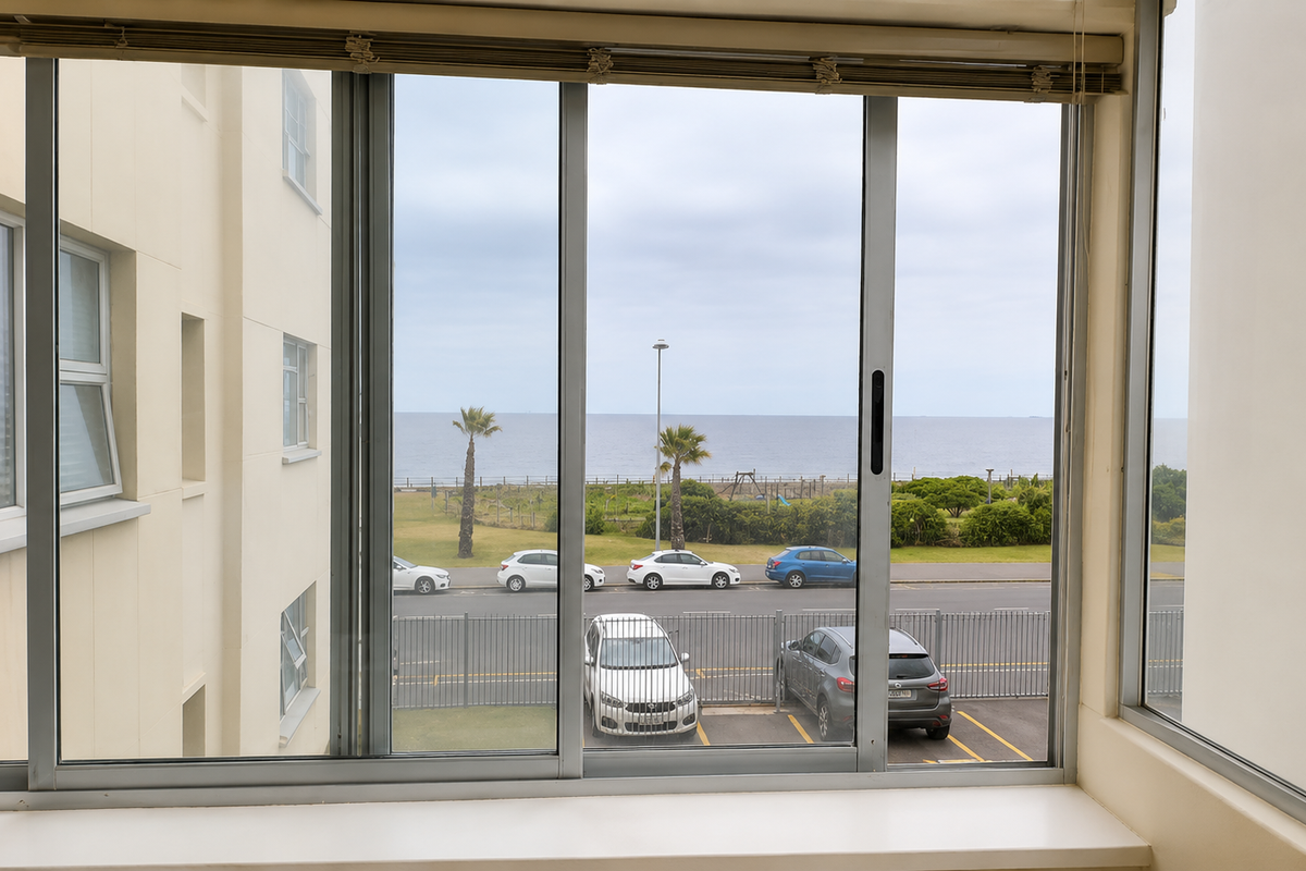 A clear view of the ocean and coastal road from an apartment window, featuring sliding aluminium frames and a clean interior sill.