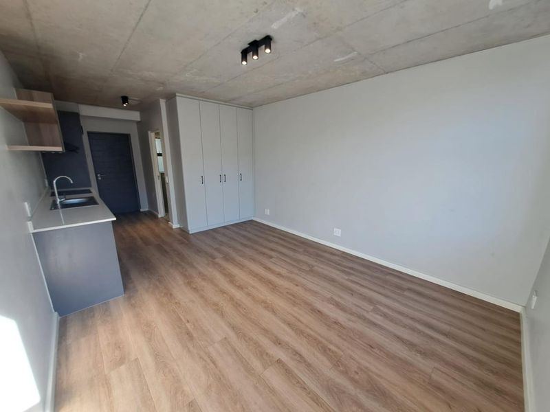 Modern open-plan living area featuring an exposed concrete ceiling, laminate flooring, built-in cupboards, and a compact kitchen with sleek cabinetry.