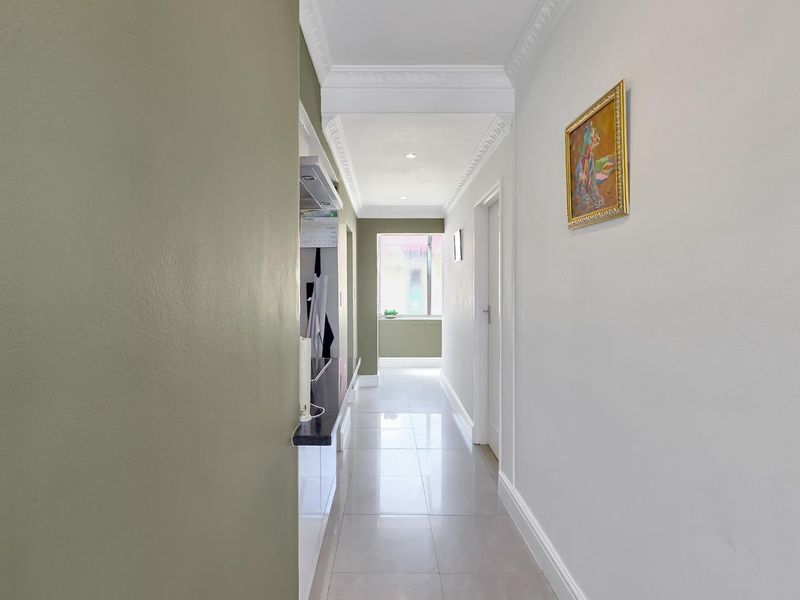 A bright and well-maintained hallway featuring tiled flooring, decorative crown molding, and recessed ceiling lights, leading towards a kitchen area.