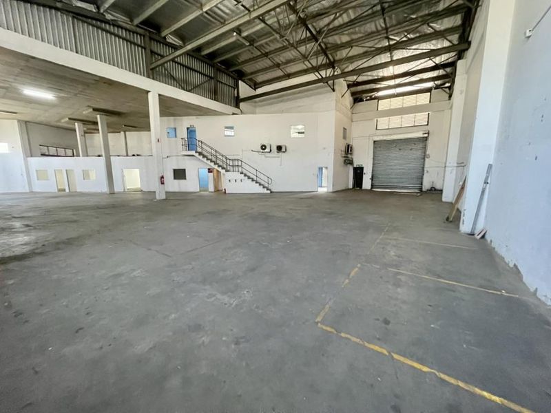 Spacious industrial warehouse featuring high ceilings with exposed steel trusses, a mezzanine level, concrete flooring, and a large roller shutter door for easy access.
