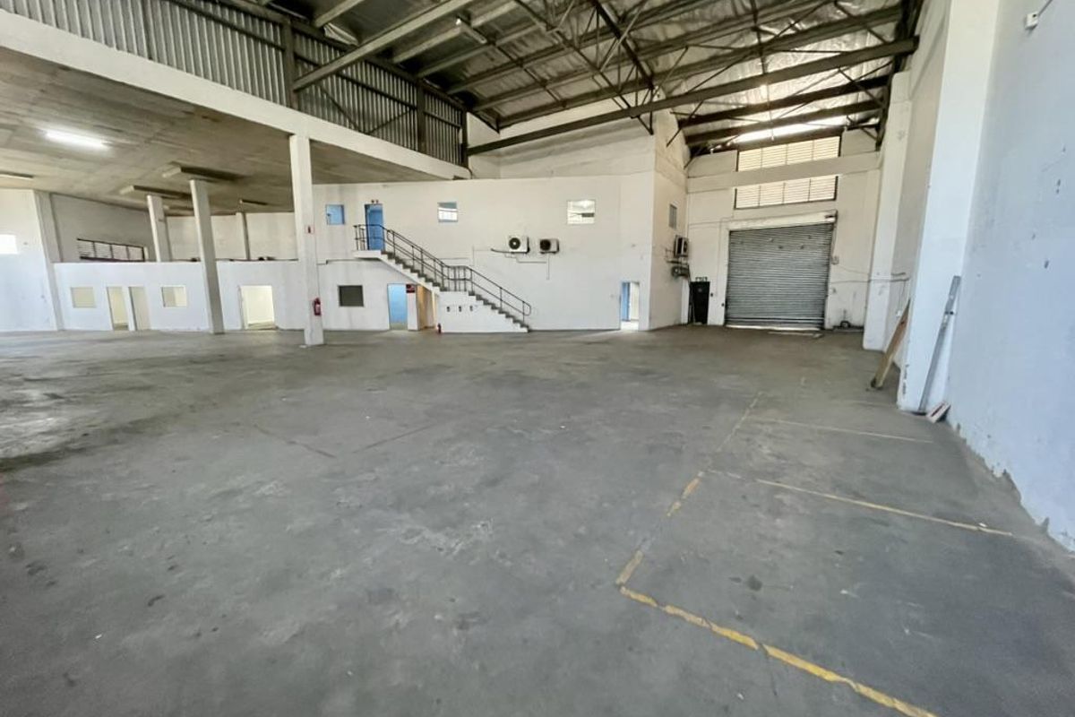 Spacious industrial warehouse featuring high ceilings with exposed steel trusses, a mezzanine level, concrete flooring, and a large roller shutter door for easy access.