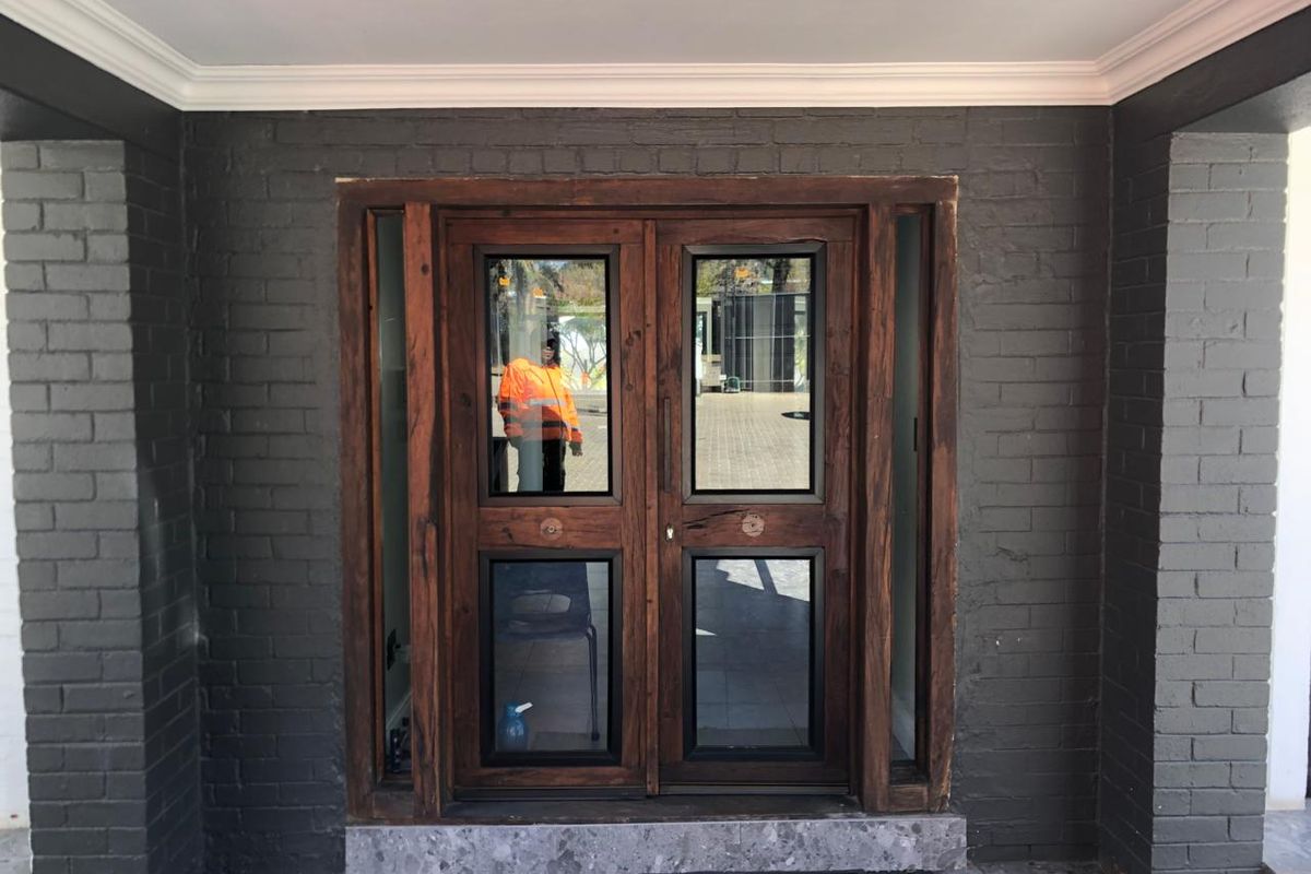 A modern office entrance featuring double timber doors with glass panels, set within a dark painted brick facade and stone threshold.