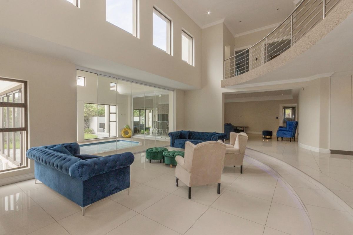 Spacious double-volume living area featuring polished tile flooring, a mezzanine level with a modern stainless steel balustrade, and a glass-enclosed indoor pool view.