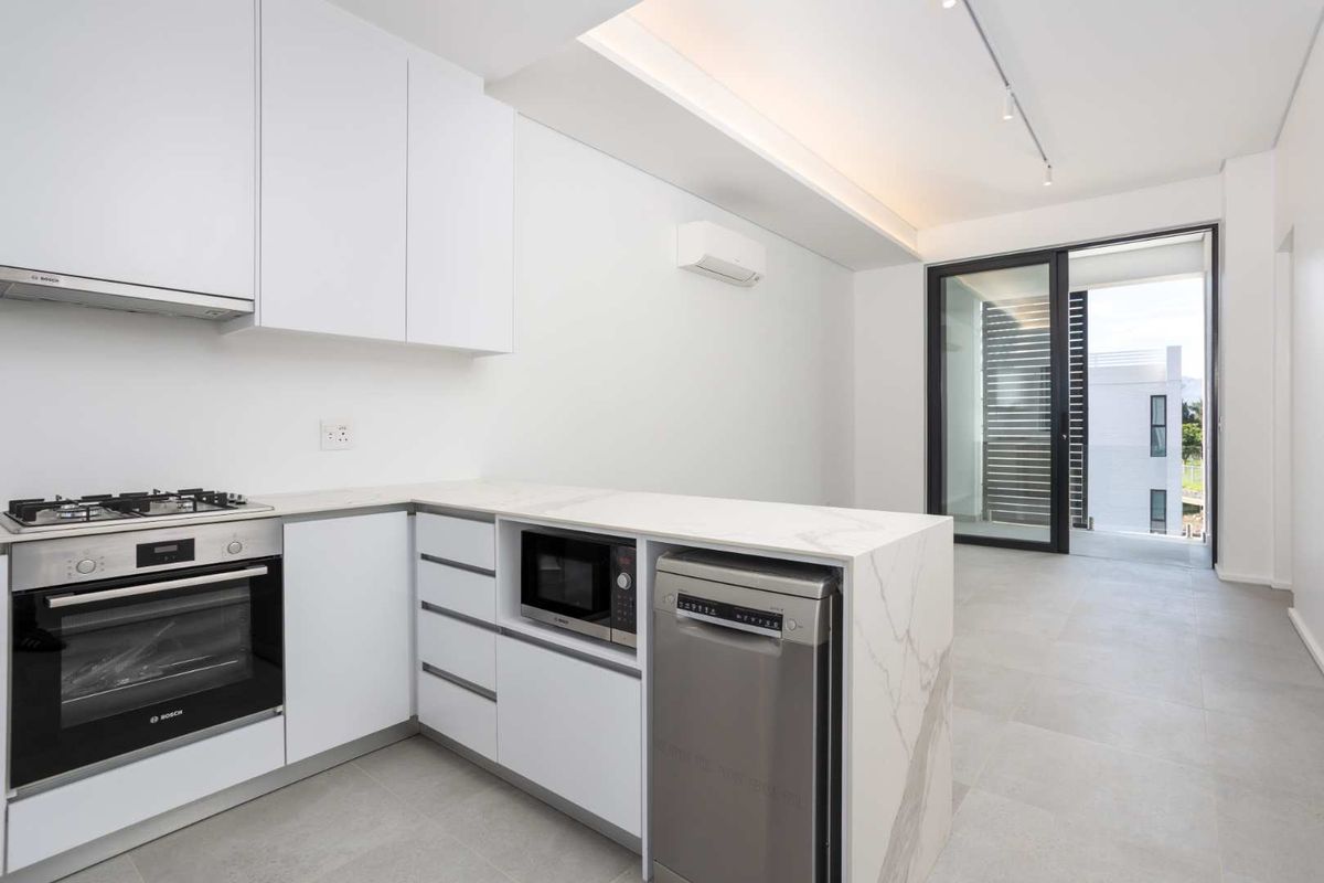 Modern open-plan kitchen featuring sleek white cabinetry, stone countertops, integrated appliances, and direct access to a private balcony through sliding glass doors.