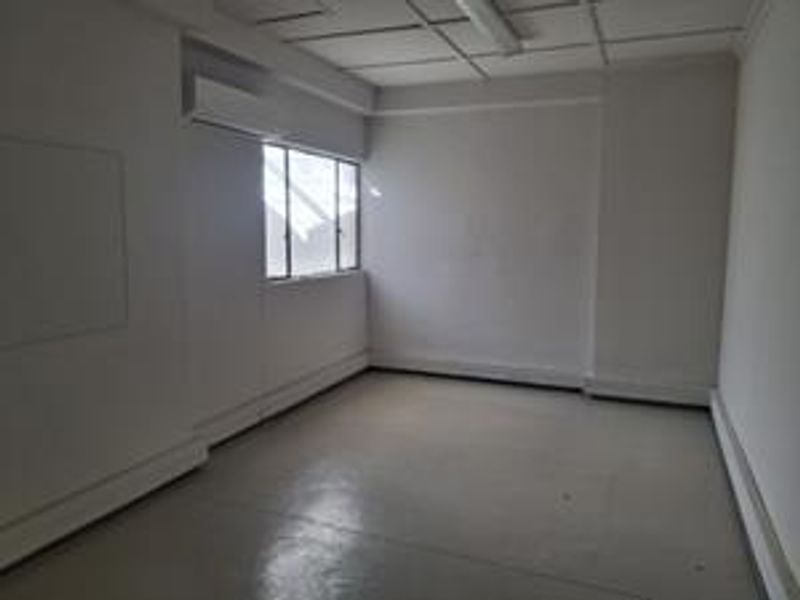 A vacant, neutral-toned office space featuring a suspended ceiling, wall-mounted air conditioning unit, and natural light from a multi-pane window.