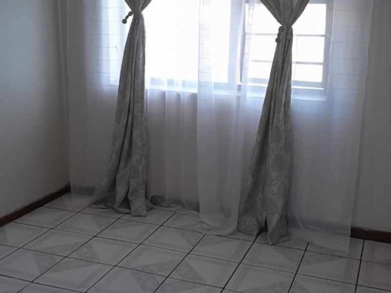 A bright, empty bedroom featuring neutral tiled flooring and a window fitted with security bars.