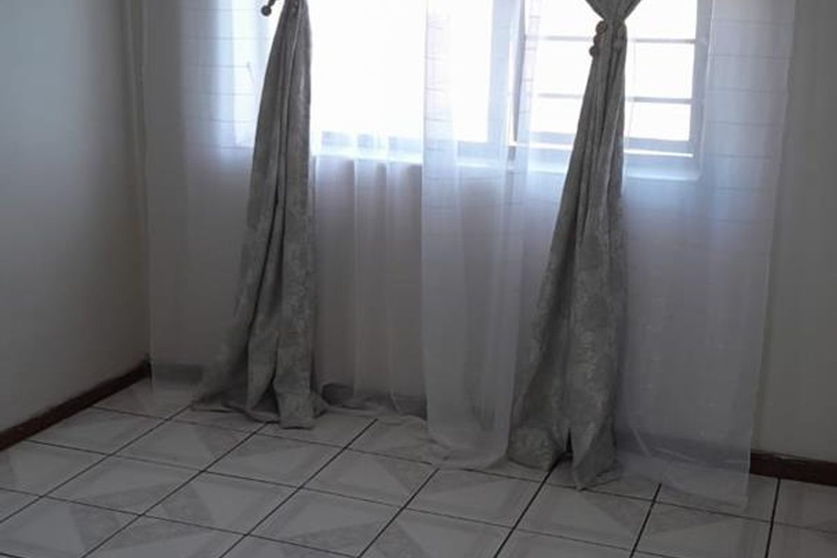 A bright, empty bedroom featuring neutral tiled flooring and a window fitted with security bars.