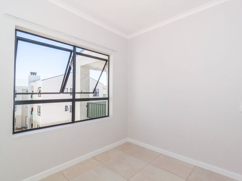 A bright, empty bedroom featuring neutral tiled flooring and a large aluminium-framed window with a view of the surrounding apartment complex.