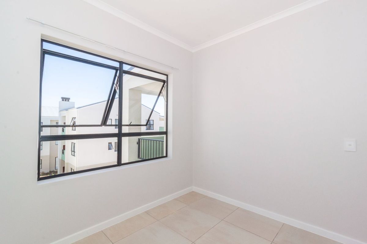 A bright, empty bedroom featuring neutral tiled flooring and a large aluminium-framed window with a view of the surrounding apartment complex.