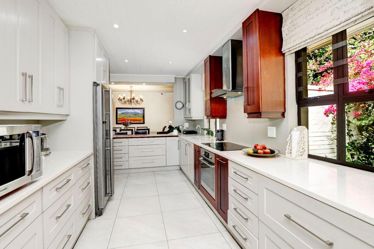 Modern galley-style kitchen featuring white cabinetry, stone countertops, built-in oven, glass cooktop, and stainless steel extractor fan, leading into an open-plan dining area.
