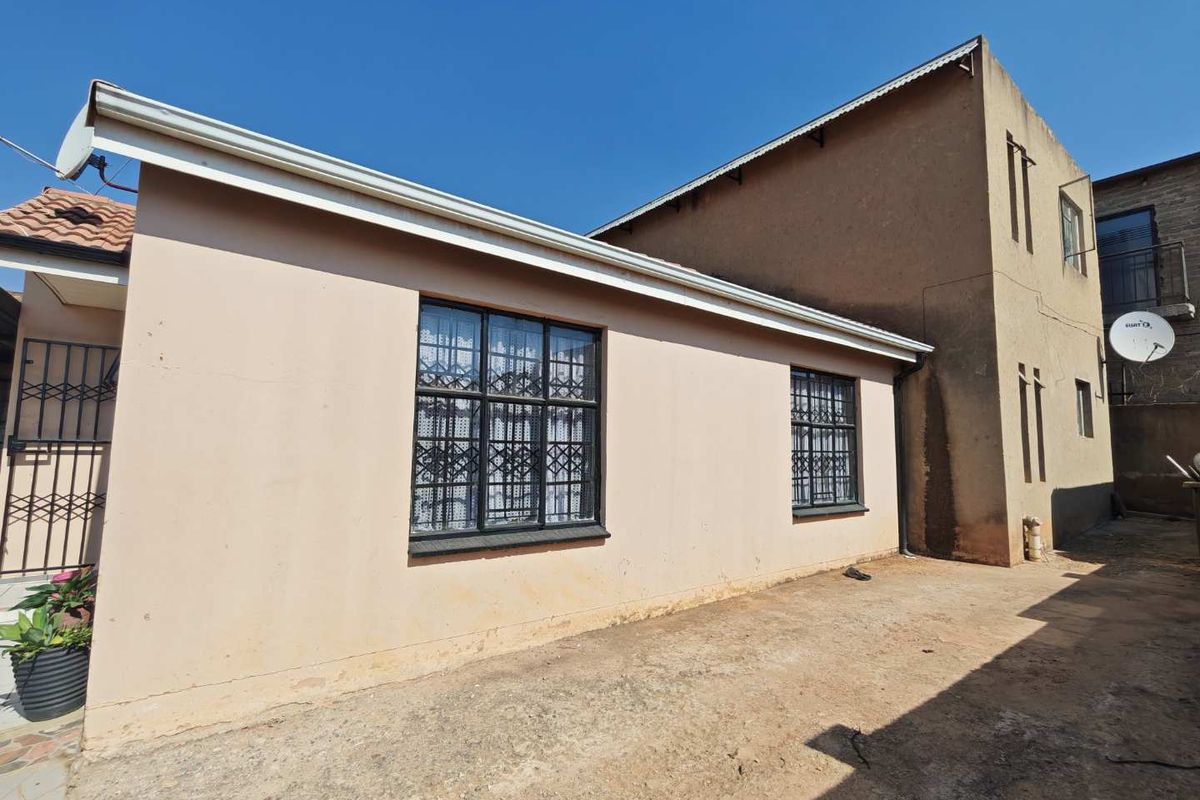 Rear exterior view of the property featuring plastered walls, security bars on windows, and an unpaved ground area.