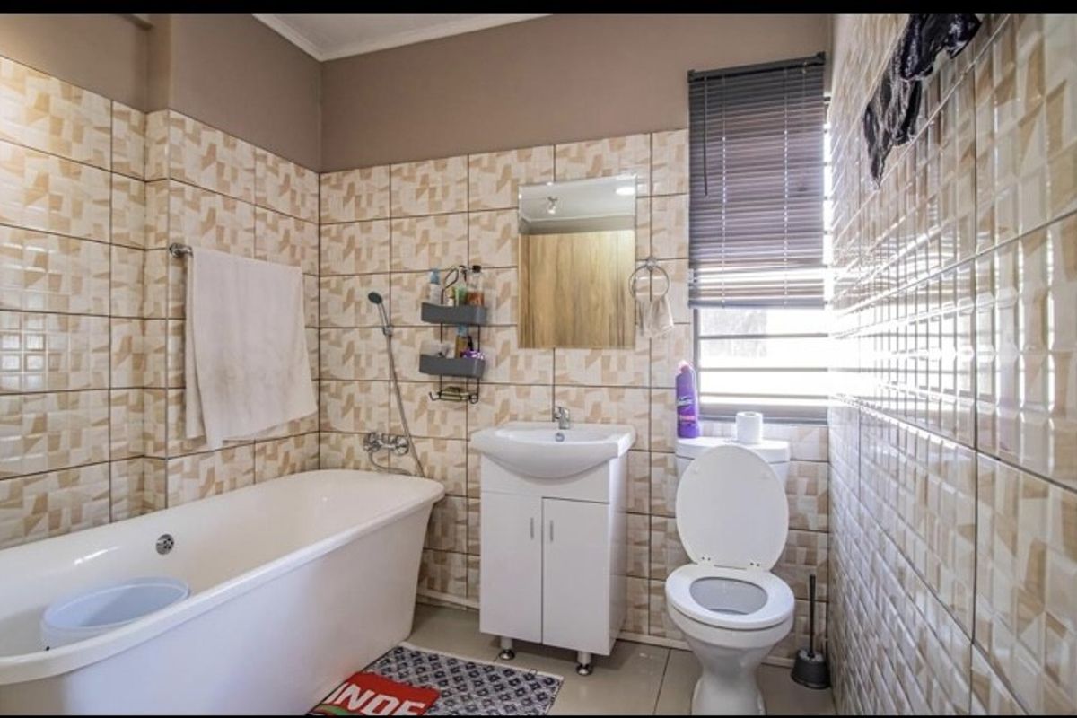 A functional bathroom featuring a bathtub, vanity basin, and toilet, finished with neutral wall tiling and a window with fitted blinds.