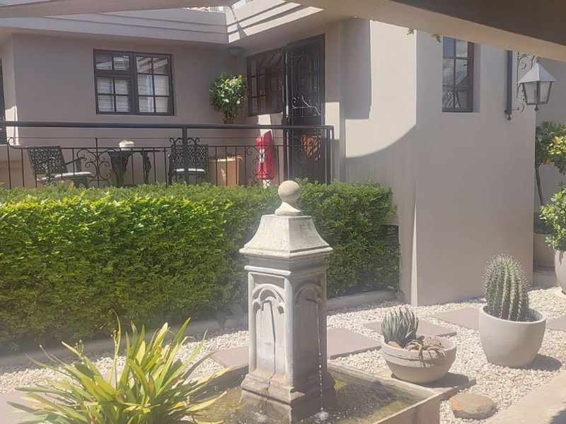 A well-maintained rear courtyard featuring a stone fountain, gravel landscaping, a manicured hedge, and a raised patio area with wrought iron railings.
