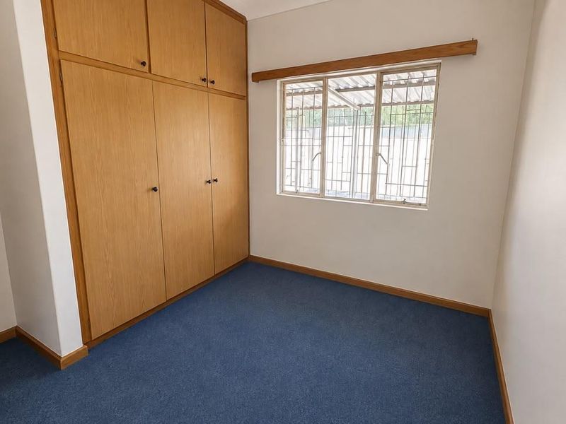 A clean, empty bedroom featuring built-in wooden cupboards, blue carpeting, and a window fitted with security bars.