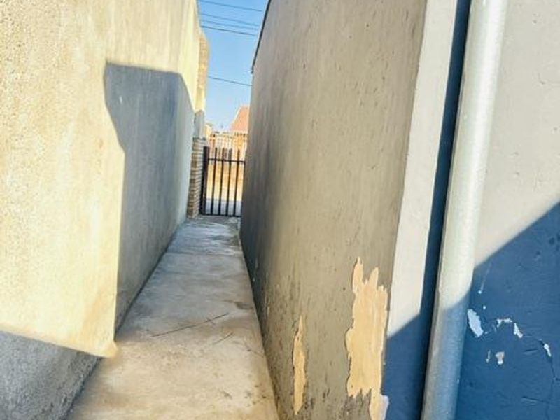 A narrow side passage between two boundary walls featuring a concrete pathway and a metal gate at the end.