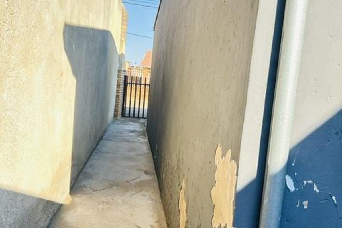 A narrow side passage between two boundary walls featuring a concrete pathway and a metal gate at the end.