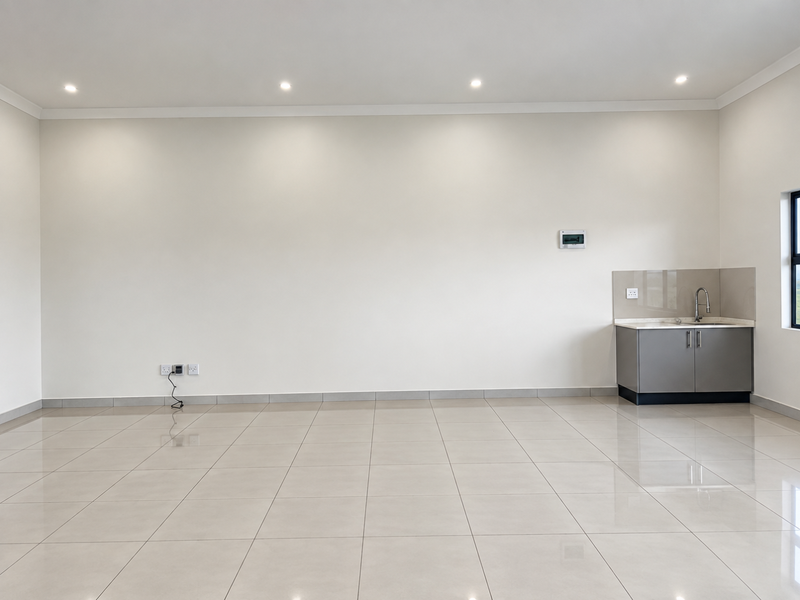 A bright, spacious open-plan area featuring neutral tiled flooring, recessed downlighting, and a compact kitchenette with built-in cabinetry and sink.