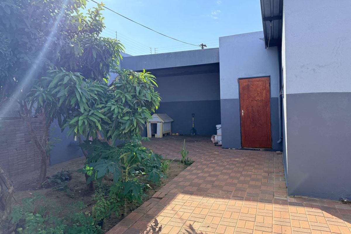 A paved rear courtyard area featuring a covered section, mature trees, and direct access to the main dwelling via a wooden door.
