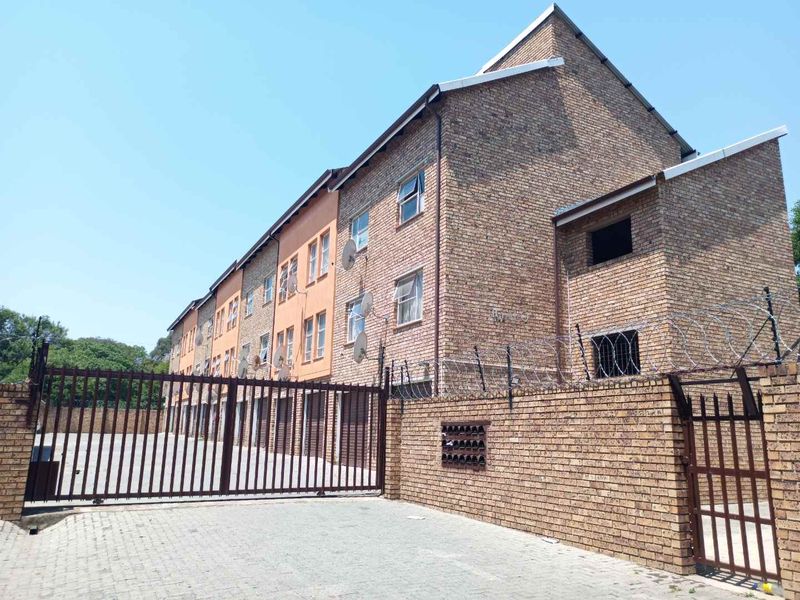 Exterior view of a multi-story apartment complex featuring face brick construction, secure gated access, paved driveway, and perimeter security fencing.