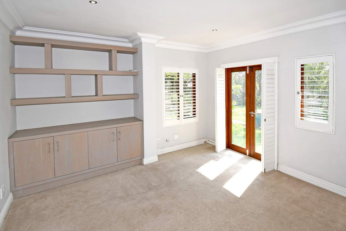 A bright, neutral living room featuring built-in cabinetry, shelving, carpeted flooring, and double doors opening to the garden with plantation shutters.