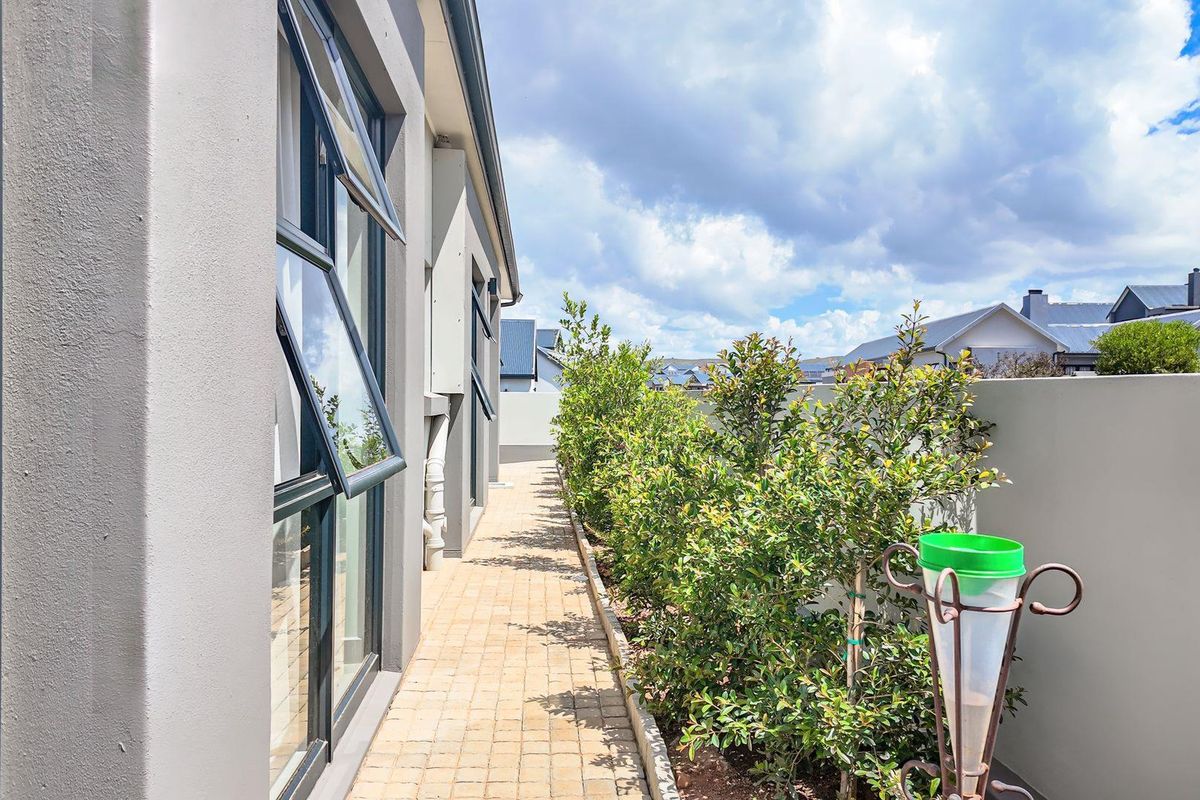 A side exterior walkway featuring paved flooring, modern aluminium window frames, and a boundary wall with established garden greenery.