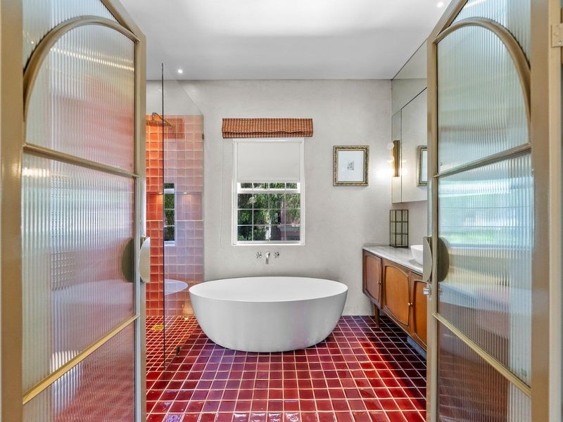 A stylish bathroom featuring a freestanding bathtub, a walk-in shower with vibrant tiled accents, and a wooden vanity, accessed through elegant reeded glass double doors.