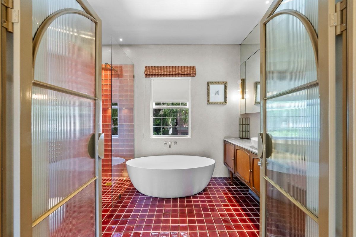 A stylish bathroom featuring a freestanding bathtub, a walk-in shower with vibrant tiled accents, and a wooden vanity, accessed through elegant reeded glass double doors.