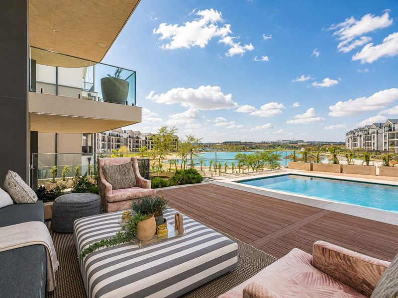 Spacious covered balcony featuring timber decking and glass balustrades, overlooking a private swimming pool and scenic estate lagoon views.