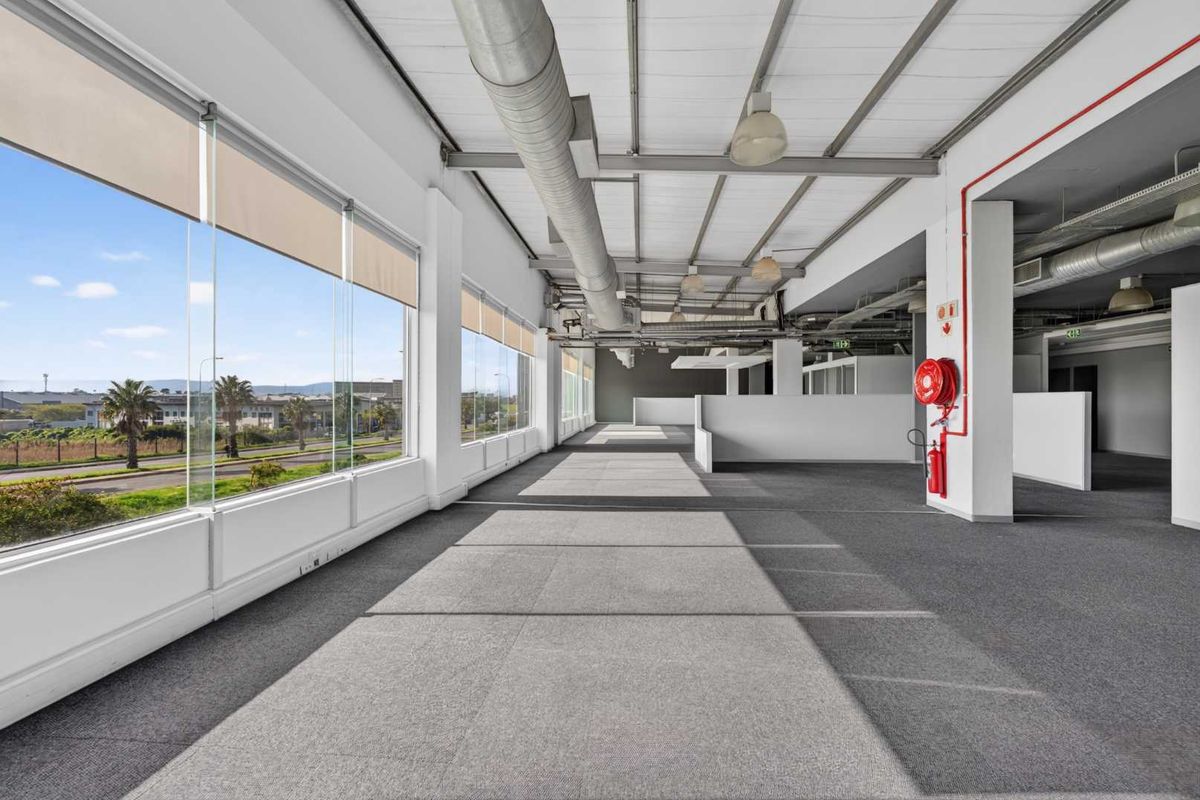 Spacious open-plan office area featuring exposed industrial ducting, carpeted flooring, and large windows providing ample natural light and exterior views.