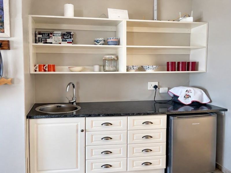 A compact scullery or kitchenette area featuring white cabinetry, granite countertops, a stainless steel sink, and open wall shelving.
