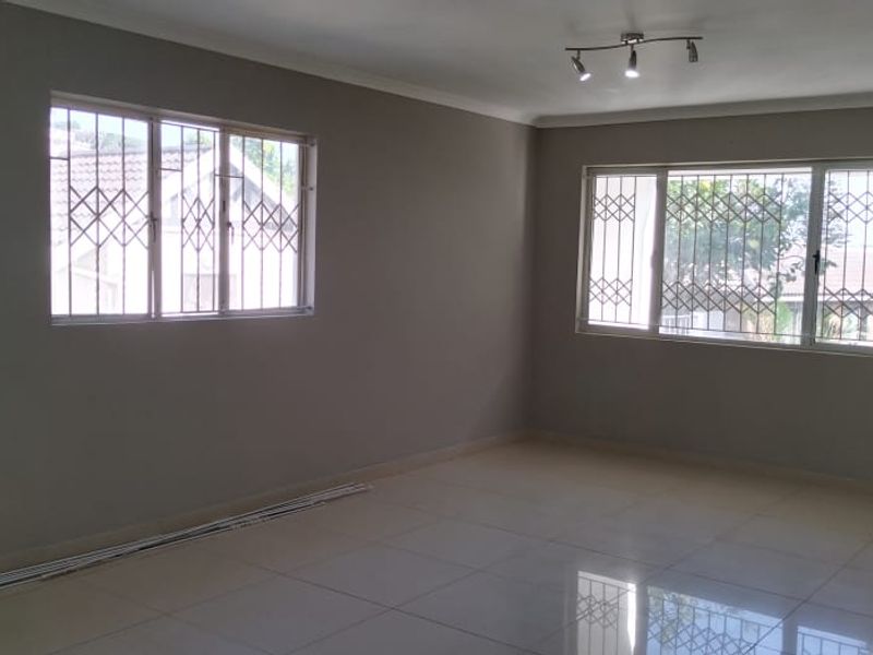 A spacious, empty living area featuring neutral grey walls, large windows with security bars, tiled flooring, and a timber entrance door.