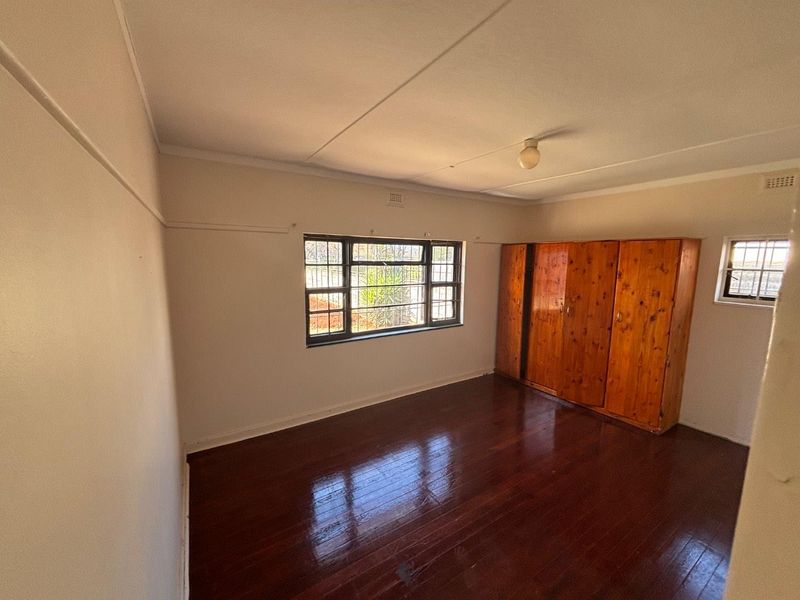 A spacious bedroom featuring polished timber flooring, built-in wooden cupboards, and steel-framed windows with security bars.