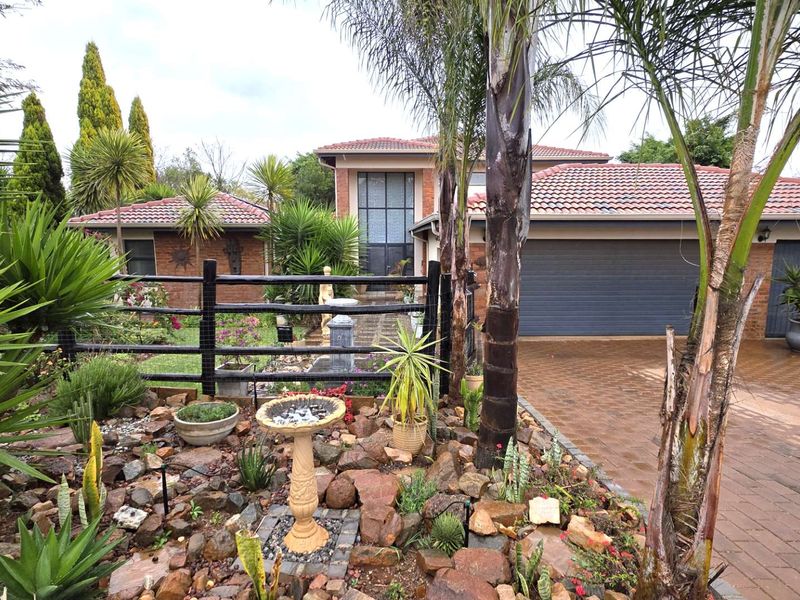 Front elevation of a brick house featuring a paved driveway, double garage, and a well-maintained landscaped garden with mature palm trees.