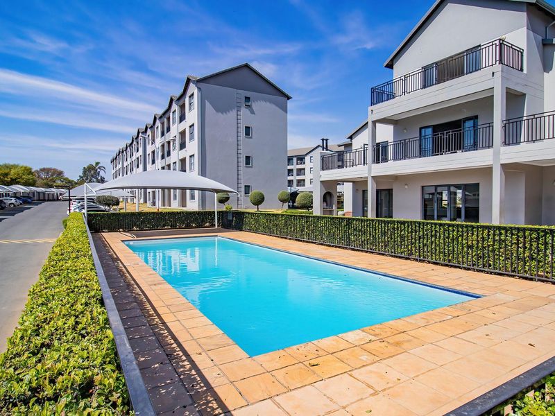 A well-maintained communal swimming pool area featuring paved surrounds, metal fencing, and shade sails, situated within a modern apartment complex.