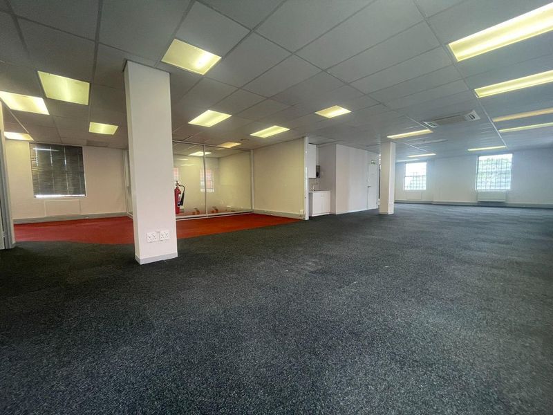 Spacious open-plan office area featuring a suspended ceiling, recessed lighting, glass-partitioned room, and a small built-in kitchenette unit.