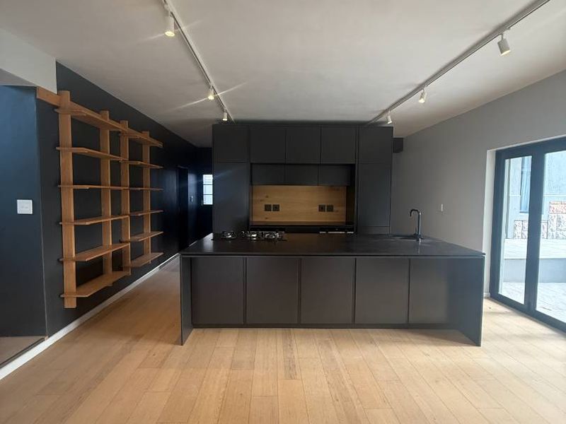 Modern, minimalist kitchen featuring a large central island, sleek dark cabinetry, integrated hob, and light timber flooring with contemporary track lighting.
