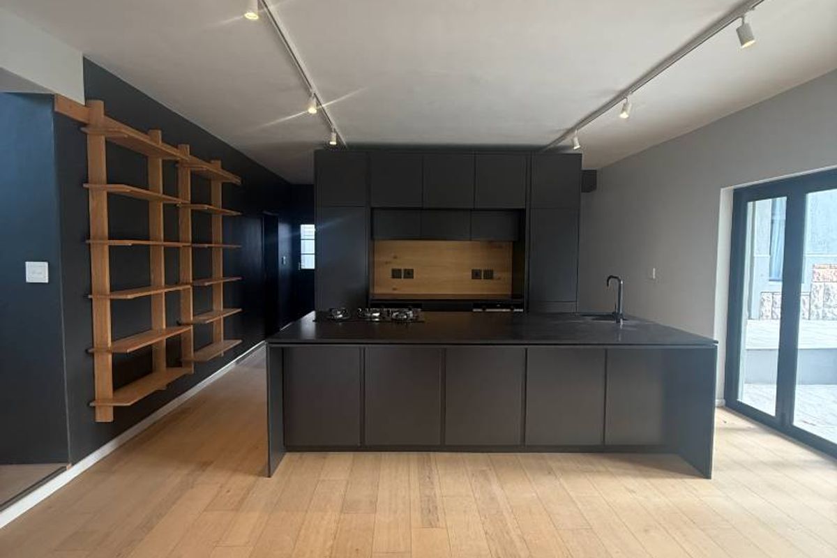 Modern, minimalist kitchen featuring a large central island, sleek dark cabinetry, integrated hob, and light timber flooring with contemporary track lighting.