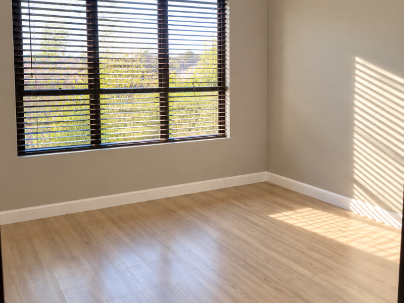 A bright, empty bedroom featuring light-toned laminate flooring, neutral wall paint, crown molding, and dark venetian blinds.